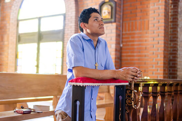 A young Latino man kneeling with a rosary in his hand, looking upward, radiating peace and...