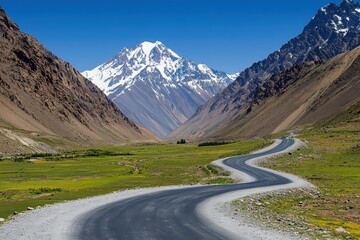 Fototapeta premium Winding mountain road through green valley, snow-capped peaks in background. Ideal for travel, adventure, and nature themes.