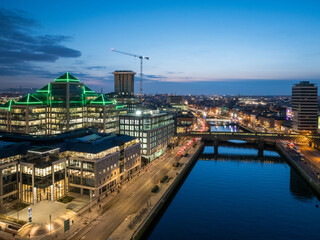 A dusk view of Dublin's modern skyline with the iconic green-lit Bank of Ireland building, a river reflecting the city lights, and a bridge crossing over it.