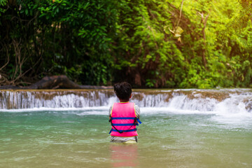 Fototapeta premium back asian child standing in waterfall or kid girl wearing life jacket to people playing stream water on nature in forest wild garden with trees on summer holiday travel at huai mae khamin waterfall
