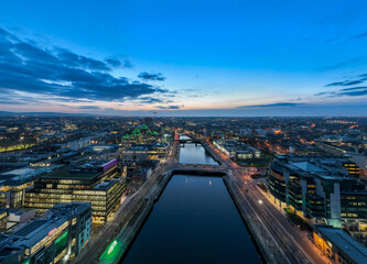A dusk view of Dublin's modern skyline with the iconic green-lit Bank of Ireland building, a river reflecting the city lights, and a bridge crossing over it.