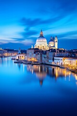 Fototapeta premium Venice cityscape at twilight, with iconic domed cathedral illuminated against vivid blue sky. Calm water reflects glowing lights and historic architecture, creating serene elegance.