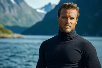 Fototapeta premium A confident man stands near the serene waters of the Norwegian Fjords, showcasing his rugged charm against a breathtaking natural backdrop of mountains and sea.