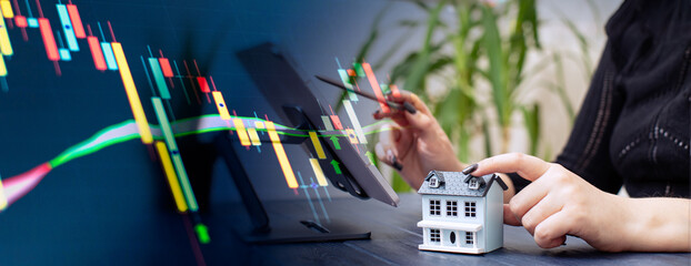Professionals Reviewing Data with Miniature House Model and Stock Market Candlestick Chart Overlay in a Business Setting