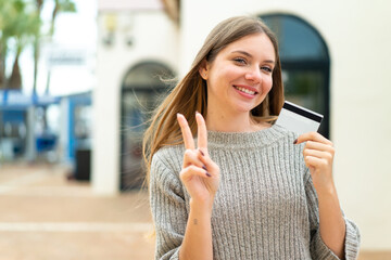 Young pretty blonde woman holding a credit card at outdoors smiling and showing victory sign