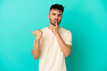 Young caucasian man isolated on blue background pointing to the side and doing silence gesture