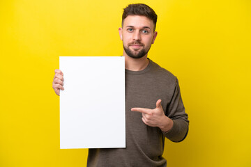 Young caucasian man isolated on yellow background holding an empty placard with happy expression and pointing it