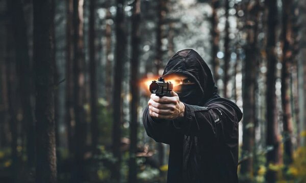 Personne encapuchonn&eacute;e tirant avec un pistolet dans une for&ecirc;t sombre, sc&egrave;ne d&rsquo;action en ext&eacute;rieur avec arme de poing, &eacute;clair de tir, capuche noire, danger, violence, crime et tension

