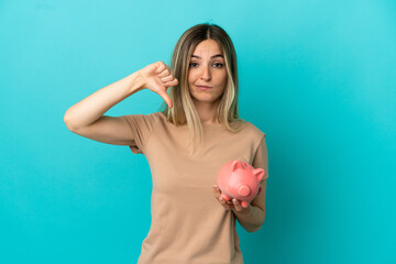 Young woman holding a piggybank over isolated blue background showing thumb down with negative...