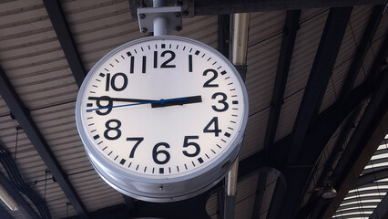 A large white analog clock hangs from the ceiling at the train station.