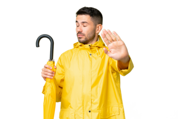 Young caucasian man with rainproof coat and umbrella over isolated background making stop gesture and disappointed