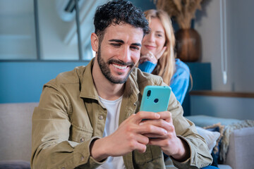 Smiling man using smartphone with a woman in the background at home