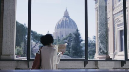 Woman tourist holds a map while admiring the view of Vatican City cathedral of St. Peter through a large window.