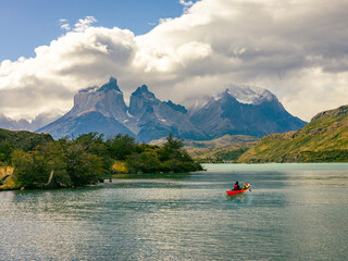 A vibrant red canoe glides across turquoise waters in Chilean Patagonia, surrounded by rugged mountains, lush greenery, and dramatic clouds in Torres del Paine.