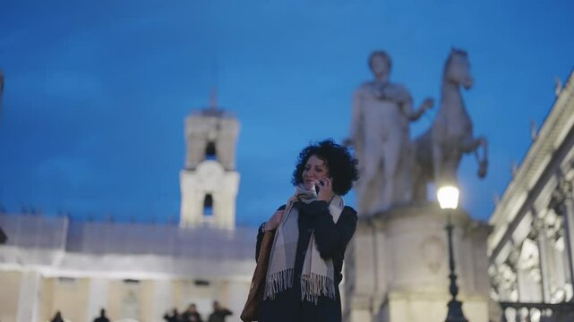 A business woman communicates talking on phone while walking at the Cordonata Capitolina with defocused Campidoglio at background in Rome during twilight, capturing the essence of work and travel.