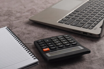 Office table desk. Workspace with calculator, notebook, laptop on dark gray background.