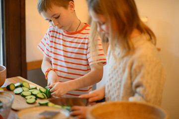 A brother and sister help their mother in the kitchen, chopping mushrooms, cucumbers, and tomatoes....