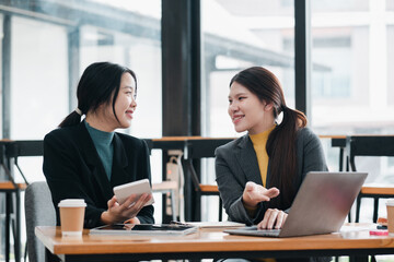 Two women in business attire discussing work at a table with a laptop and smartphone in a bright, modern office setting.