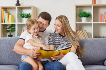 Young happy family sitting on couch at home, reading book to their daughter. Smiling little girl...