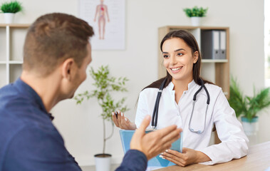Friendly female physician or cardiologist sitting at desk in office, listening to patient...