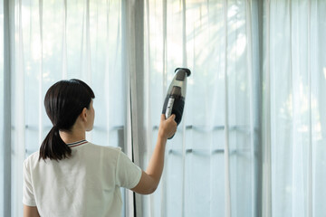 Young maid using handheld cordless vacuum cleaner to vacuuming and cleaning the dust on curtains in...