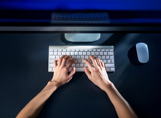 Fototapeta premium Overhead view of hands typing on a white keyboard in dimly lit room at night. A computer monitor and mouse are visible on a dark surface.