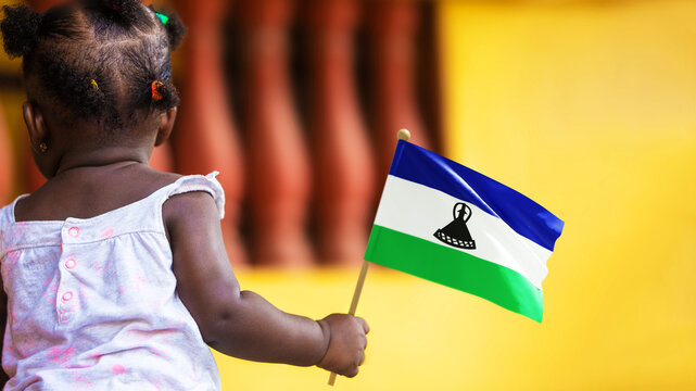 Adorable black African kid female baby todler girl standing holding Lesotho national flag in hand. October 4 Independence Day celebration. back view
