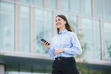 Fototapeta premium Young woman walking outside while using smartphone near modern building in bright daylight