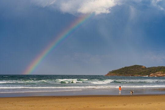 distant view of a woman walking a dog on a beach with a colourful rainbow overhead in a dark sky