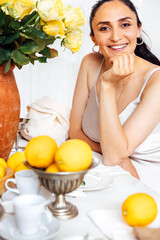 Obraz premium Young woman is sitting and laughing at table. Yellow roses, kitchen utensils and lemons in fruit vase. Joyful girl in white satin dress. Vintage interior.