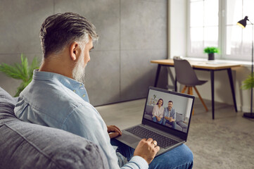 Side view shot of businessman making video call to his friends. Gray haired bearded middle aged man sitting on couch with laptop on his laps engaged in webcam conference conversation with relatives
