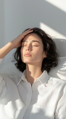 Fototapeta premium A serene portrait of a woman with closed eyes, resting gently with one hand on her forehead, bathed in soft natural light.