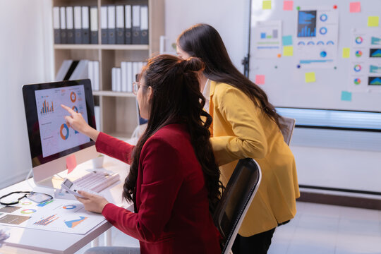 Two businesswomen are collaborating on a project, analyzing financial charts displayed on a computer screen in a modern office setting, pointing at the monitor and discussing data