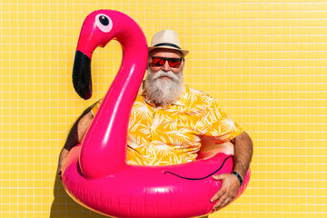 Happy funny senior man portrait on yellow wall with flamingo air mat during summertime © oneinchpunch