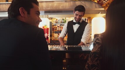 Focusing smiling smart bartender serving gin fizz or tonic soda at luxurious bar, engaging lively conversation with VIP customer couple at back view in atmosphere at lighting warm nightclub. Vinosity.