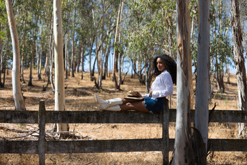 Fototapeta premium woman, young, beautiful, Latin, brunette with curly hair dressed in white shirt, denim skirt, white boots and hat, sitting with her legs stretched out on the forest fence. The woman rests.
