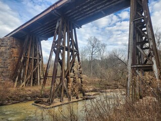 An aged railway trestle bridge standing over a serene stream, surrounded by natural vegetation.