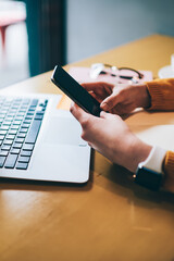 Fototapeta premium Cropped image of female hands holding smartphone device with good internet connection sitting with laptop computer at coffee shop, woman searching information on cellular phone for planning trip