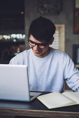 Cheerful male graphic designer holding modern telephone in hand while sitting at digital laptop computer connecting to internet and notepad with blank screen area for your advertising text