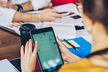 Cropped back view of young woman holding modern touch pad device in hands and searching app on internet website using wireless 4G connection during working process with colleagues in office
