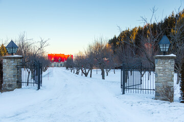 Naklejka premium Traditional stone house with steep red metal roof seen at the end of an alley covered in fresh snow during a winter morning, Saint-Nicolas, Quebec, Canada