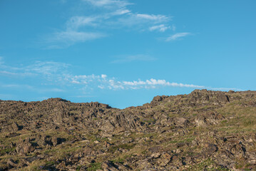 Nature layered background with sunlit grassy rocky hill under clouds in blue sky. Minimalist view to stone hill in bright sun in changeable weather. Natural backdrop of rock mountain pass in sunny day