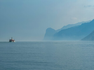 Ferry boat floating along coastline of misty Lake Garda covered with haze seen from Nago-Torbole, Trentino, Northern Italy. Steep rugged cliffs and mountain peaks, Garda Prealps. Dramatic cloudy sky