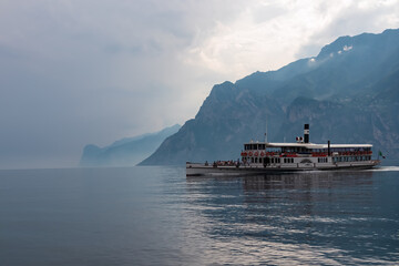 Ferry boat floating along coastline of misty Lake Garda covered with haze seen from Nago-Torbole, Trentino, Northern Italy. Steep rugged cliffs and mountain peaks, Garda Prealps. Dramatic cloudy sky