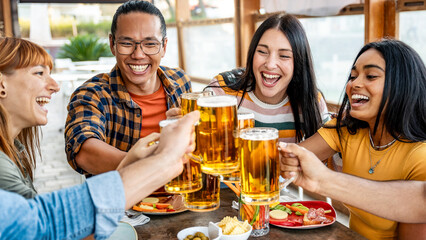 Happy young people enjoying happy hour sitting at bar restaurant - Multiracial friends toasting beer glasses at brewery pub patio - Friendship concept with guys and girls drinking and eating together