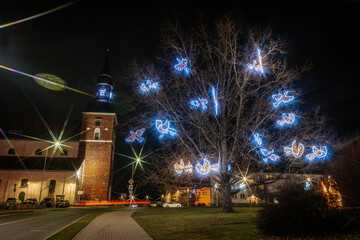 A glowing tree adorned with blue-lit doves near a historic brick church at night. Vibrant...