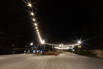 Fototapeta premium A serene park at night featuring a pathway lit by decorative string lights and lampposts. Trees and landscaped areas enhance the tranquil atmosphere.