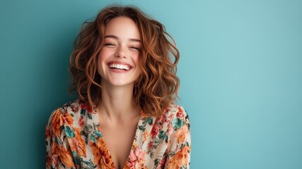 A woman with brown hair bursts into laughter, radiating joy and warmth, wearing a lovely floral patterned outfit against a bright teal backdrop.