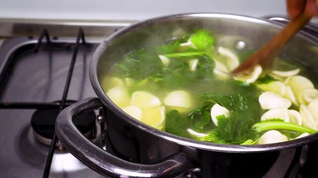Turnip greens and orecchiette are being boiled together in a stainless steel pot on a gas stove as the traditional recipe of orecchiette con le cime di rape, an italian traditional dish, requires