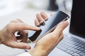 Man engaged with his modern smartphone, laptop slightly out of focus nearby against the blurred...